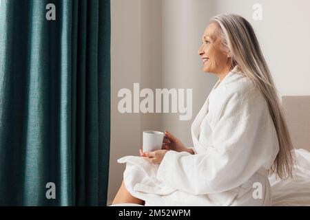 Femme âgée assise sur le lit dans la chambre et vue loin. Vue latérale de la femelle mature pensive. Banque D'Images