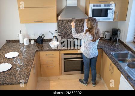 Femme blanche senior qui cuisine chez elle et touche les commandes de la hotte aspirante de la cuisine. Banque D'Images