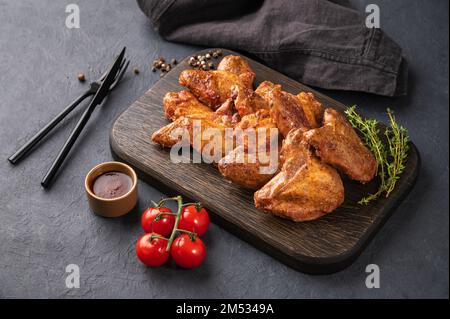 Ailes de poulet grillées avec sauce barbecue, tomate et herbe fraîche sur un panneau de bois sur fond sombre. Banque D'Images