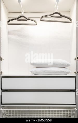 Intérieur d'un placard ouvert avec portes blanches, porte-chemises, oreillers blancs, tiroirs et panier en fil de fer Banque D'Images
