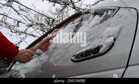 une personne dans une veste rouge nettoie la fenêtre d'une voiture grise des dérives de neige avec une brosse avec une poignée en bois et des poils roses. vue de dessous Banque D'Images