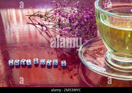Goiania, Goiás, Brésil – 25 décembre 2022: Détail d'une tasse de verre avec thé, sur la table brillante, avec le mot 'anxiété' écrit avec des pièces et des fleurs Banque D'Images