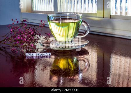 Goiania, Goiás, Brésil – 25 décembre 2022: Détail d'une tasse de verre avec thé, sur la table brillante, avec le mot 'anxiété' écrit avec des pièces et des fleurs Banque D'Images
