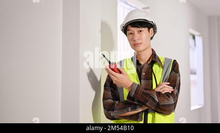 Jeune ingénieur de construction asiatique élégant et élégant, en casque rigide uniforme et blanc, tenant un talkie-walkie. Banque D'Images