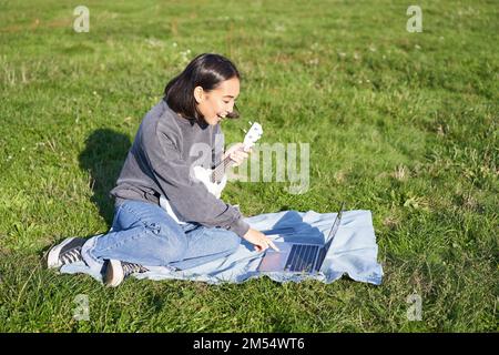 Bonne fille asiatique souriante appréciant le printemps, jouant ukulele dans le parc, à la recherche d'accords en ligne sur ordinateur portable, apprenant la nouvelle chanson Banque D'Images