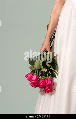 Anonyme femme en robe blanche avec bouquet de roses rouges vives debout sur fond gris le jour du mariage Banque D'Images