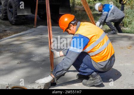 Le Slinger pose une dalle de béton sur le chantier de construction le jour de l'été. Le travailleur dans la veste de protection et le casque de construction supervise la pose de la base sur le chantier. Banque D'Images