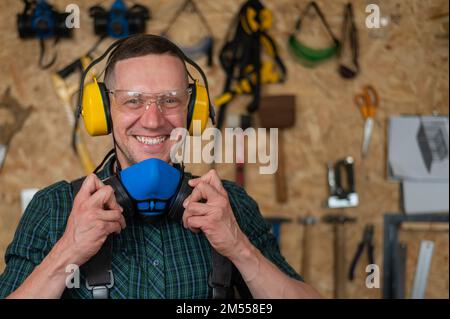 Un menuisier dans des lunettes et des couvre-oreilles enlève un respirateur de protection. Banque D'Images