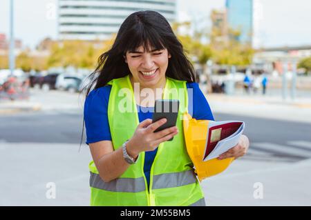 jeune femme latine caucasienne ingénieur debout dans la rue en souriant lisant les messages texte et les notifications sur les médias sociaux, l'industrie et la technolo Banque D'Images