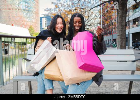 Deux jeunes femmes multiraciales souriant et regardant à l'intérieur d'un sac de shopping assis sur un banc. Quelques amis heureux montrant leurs achats sur une semaine de vente. Concept de consommables. Photo de haute qualité Banque D'Images