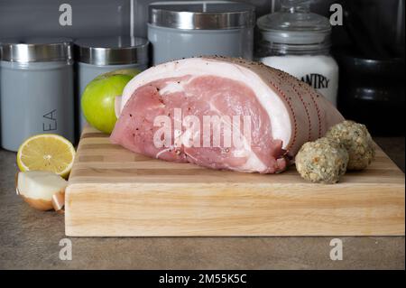 Branche de porc non cuite sur une planche à découper en bloc de hêtre avec oignon, pomme de cuisson au citron et sauge et boulettes d'oignon Banque D'Images