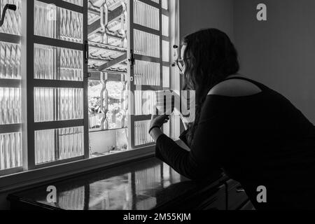 Goiania, Goiás, Brésil – 25 décembre 2022 : une femme, seule, tenant une tasse de thé et regardant la fenêtre. Image en noir et blanc. Banque D'Images