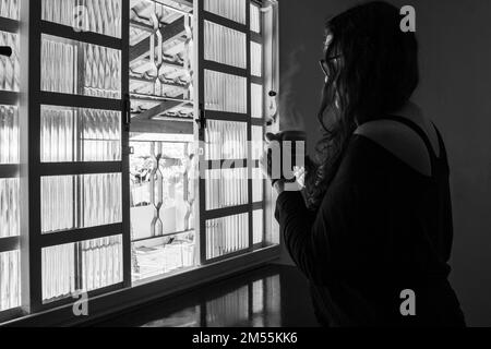 Goiania, Goiás, Brésil – 25 décembre 2022 : une femme, seule, tenant une tasse de thé et regardant la fenêtre. Image en noir et blanc. Banque D'Images