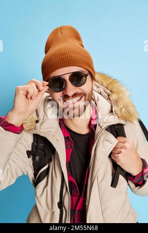 Homme mûr tendance en manteau et chapeau d'hiver avec une expression du visage heureuse regardant l'appareil photo isolé sur fond bleu. Vacances, voyage, aventure Banque D'Images