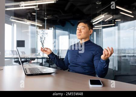 Jeune homme d'affaires asiatique fatigué méditant au travail. Il s'assoit à la table du bureau, prend une pause, ferme les yeux, les mains en position lotus, se détend. Banque D'Images