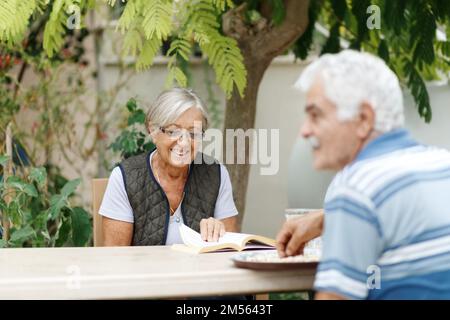 Amoureux des conjoints âgés se reposant dans le jardin et appréciant des jours, assis à l'extérieur.femme lisant le livre, homme en face d'elle. Banque D'Images