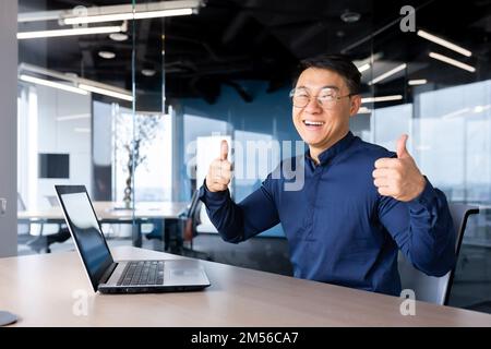 Portrait d'un jeune homme beau, un designer asiatique, un ingénieur, un pigiste. Il est assis au bureau à la table avec un ordinateur portable, regarde la caméra, souriant, montre ses mains avec ses doigts super. Banque D'Images