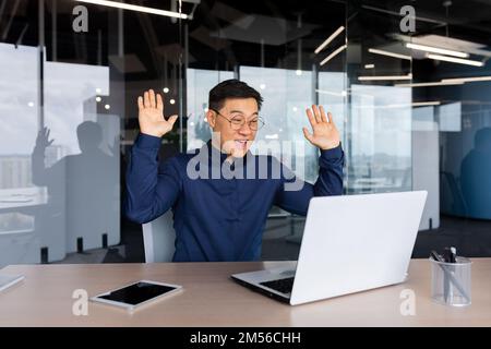 Jeune homme asiatique indépendant, homme d'affaires dans le bureau à la table parlant par appel vidéo avec des partenaires, réunion en ligne, se réjouit, félicite, a levé la main. Banque D'Images