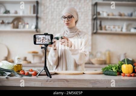 Femme moderne blogger tenant concombre en direct streaming et enregistrement vidéo recette pour la nourriture blog. Jeune femme musulmane dans hijab cuisant de la nourriture en ligne à la cuisine maison. Style de vie, concept de loisirs à la maison. Banque D'Images