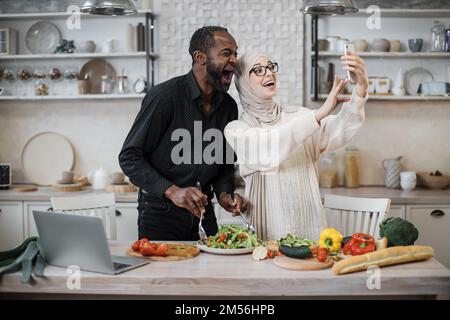 Mignon couple multinational prenant le selfie ou d'avoir la conversation vidéo avec des amis tout en ayant un petit déjeuner sain à la maison, en utilisant le tout nouveau téléphone mobile, intérieur de cuisine, espace de copie Banque D'Images