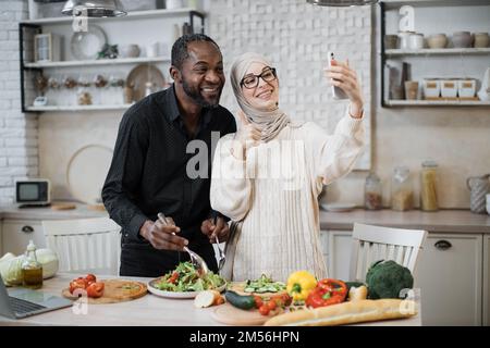 Homme africain et femme musulmane souriant et se montrant à la main tout en effectuant un appel vidéo sur smartphone et en cuisant de délicieuses salades de légumes frais dans une cuisine moderne. Banque D'Images