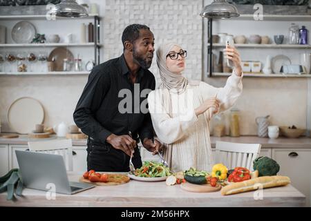 Mignon couple multinational prenant le selfie ou d'avoir la conversation vidéo avec des amis tout en ayant un petit déjeuner sain à la maison, en utilisant le tout nouveau téléphone mobile, intérieur de cuisine, espace de copie Banque D'Images