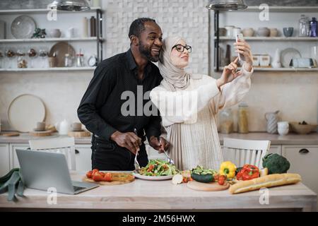 Mignon couple multinational prenant le selfie ou d'avoir la conversation vidéo avec des amis tout en ayant un petit déjeuner sain à la maison, en utilisant le tout nouveau téléphone mobile, intérieur de cuisine, espace de copie Banque D'Images