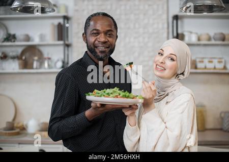 Gros plan portrait d'un couple multinational marié cuisant des aliments sains dans la cuisine, jeune femme musulmane en hijab nourrissant son mari à la fourchette avec de la salade de légumes frais. Banque D'Images