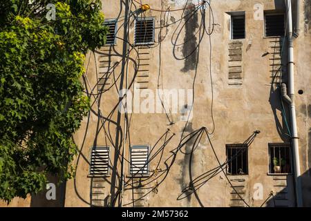 Bâtiment aux intempéries avec de petites fenêtres et des câbles électriques situés sur une rue ensoleillée près d'un arbre luxuriant Banque D'Images