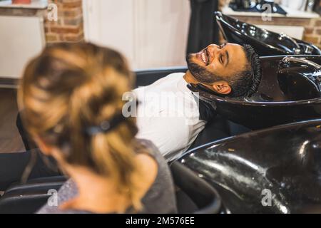 Homme en train de rire en utilisant une chaise de lavage de cheveux de salon. Tête floue d'un autre client au premier plan. Concept de coiffeur. Photo de haute qualité Banque D'Images