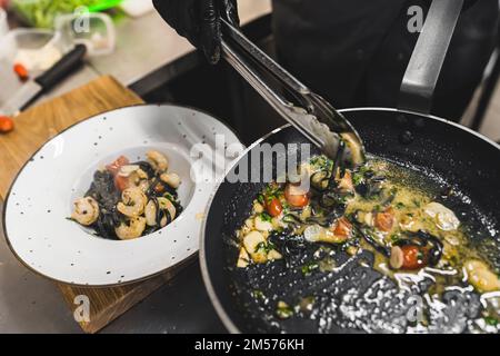 Chef complétant un plat. Vue de dessus de la personne utilisant des pinces de cuisson pour placer les pâtes de tagliolini noires avec les crevettes sur la plaque blanche. Photo de haute qualité Banque D'Images