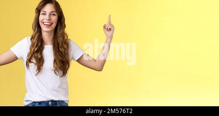 Joyeux amusé heureux animé jeune fille longue coupe de cheveux bouclés lever les mains pointant vers le haut sourire Toothy heureusement caméra présenter une excellente variante suggérer Banque D'Images