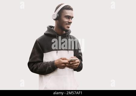 Souriant jeune homme dans des vêtements décontractés écouter de la musique et tenir son téléphone cellulaire entre les mains.Studio portrait de beau noir homme dans les écouteurs Banque D'Images