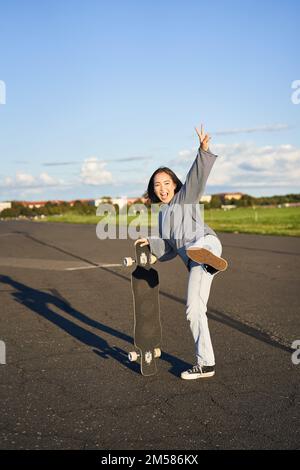 Photo verticale d'une fille de patineuse asiatique heureuse, sautant, debout avec un skateboard et souriant. Femme patinant à bord de la longboard et s'amusant Banque D'Images