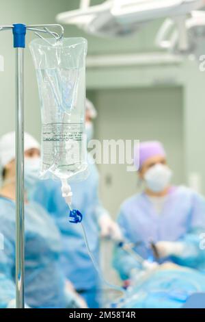 Poche avec solution de perfusion saline pour compte-gouttes dans la salle d'opération en arrière-plan de la chirurgie. Mise au point sélective. Silhouettes de chirurgiens sur un fond flou. Banque D'Images