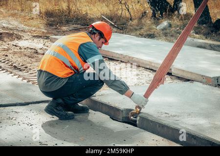 Le Slinger pose une dalle de béton sur le chantier de construction le jour de l'été. Le travailleur dans la veste de protection et le casque de construction supervise la pose de la base sur le chantier. Banque D'Images