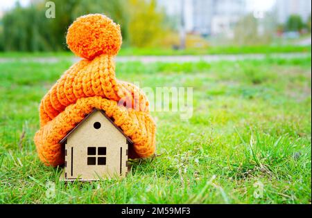 Chapeau tricoté volumineux mis sur un jouet modèle de maison en bois placé sur une pelouse verte à l'extérieur. Rester au chaud sans démarrer le chauffage. Banque D'Images