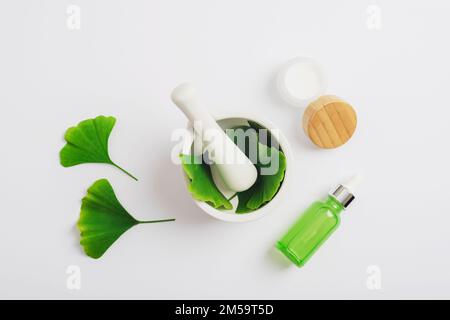 Feuilles de ginkgo vert frais, flacon de sérum cosmétique et pot à crème, mortier et pilon sur fond blanc. Concept de cosmétiques naturels faits à la main. Vue de dessus, f Banque D'Images