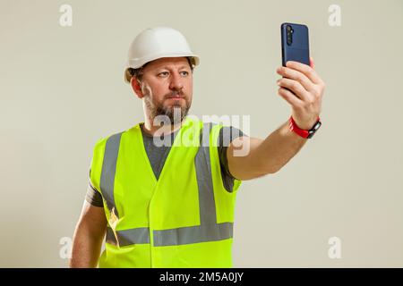 Un homme travailleur dans un gilet et un casque de construction blanc prend un selfie sur le téléphone sur un fond blanc Banque D'Images