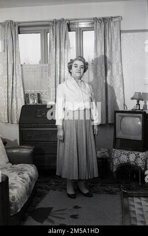 1950s, historique, une dame dans une pièce de devant d'une maison debout pour une image à côté d'un ensemble de télévision de l'époque, Angleterre, Royaume-Uni. Banque D'Images