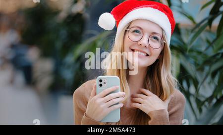 Heureux succès stupéfié femme blanche fille dame en rouge Santa Claus chapeau de Noël et de lunettes de gagner avec téléphone mobile victoire offre réalisation nouvel an Banque D'Images