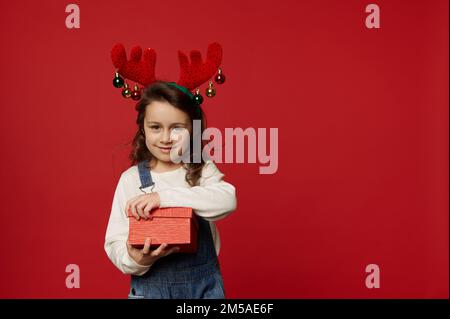 Adorable petite fille portant une combinaison en denim bleu, un pull blanc et un cerceau de cerf, déballant une boîte cadeau pour Noël Banque D'Images