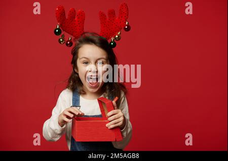 Bébé jeune fille espiègle surjoyeuse avec des cerfs et des rancune rigole à la caméra, pose avec le cadeau de Noël sur fond rouge Banque D'Images