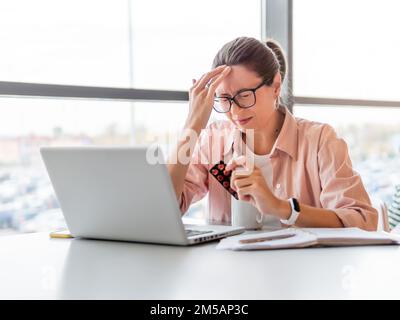 Une femme qui frognait regarde des pilules de médicaments tout en travaillant avec un ordinateur portable. Problèmes de santé mentale, épuisement émotionnel ou maux de tête. Bureau moderne au co-working c Banque D'Images