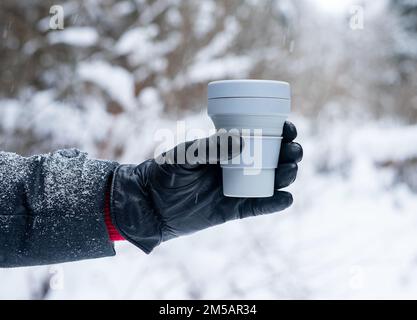 Main dans le gant tenant la tasse à café en silicone écologique à emporter, retirez la tasse à thé en hiver, par temps neigeux en plein air. Photo de haute qualité Banque D'Images