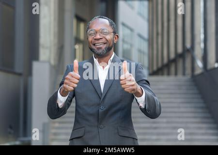 Portrait d'un homme d'affaires afro-américain mature, homme d'affaires en costume souriant et regardant l'appareil photo montrant les pouces heureux devant le bâtiment de bureau. Banque D'Images