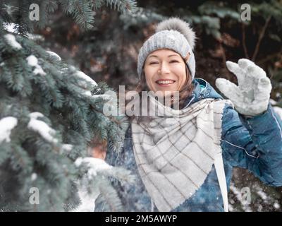 Femme souriante dans le chapeau tricoté de câble joue avec la neige. Amusez-vous dans le parc entre des sapins enneigés. Une femme rit alors qu'elle lance un ballon de neige. Saison froide. Banque D'Images