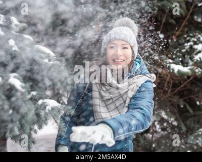 Femme souriante dans le chapeau tricoté de câble joue avec la neige. Amusez-vous dans le parc entre des sapins enneigés. Une femme rit alors qu'elle lance un ballon de neige. Saison froide. Banque D'Images