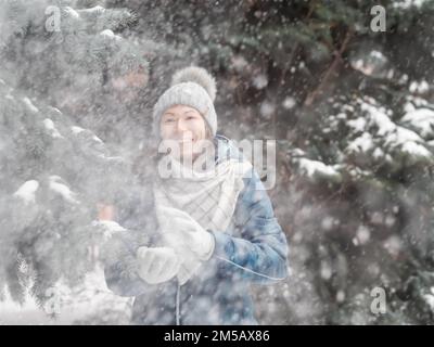 Femme souriante dans le chapeau tricoté de câble joue avec la neige. Amusez-vous dans le parc entre des sapins enneigés. Une femme rit alors qu'elle lance un ballon de neige. Saison froide. Banque D'Images