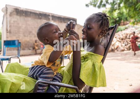 Belle fille africaine riant et jouant à thé avec son petit frère qui est assis dans ses bras Banque D'Images